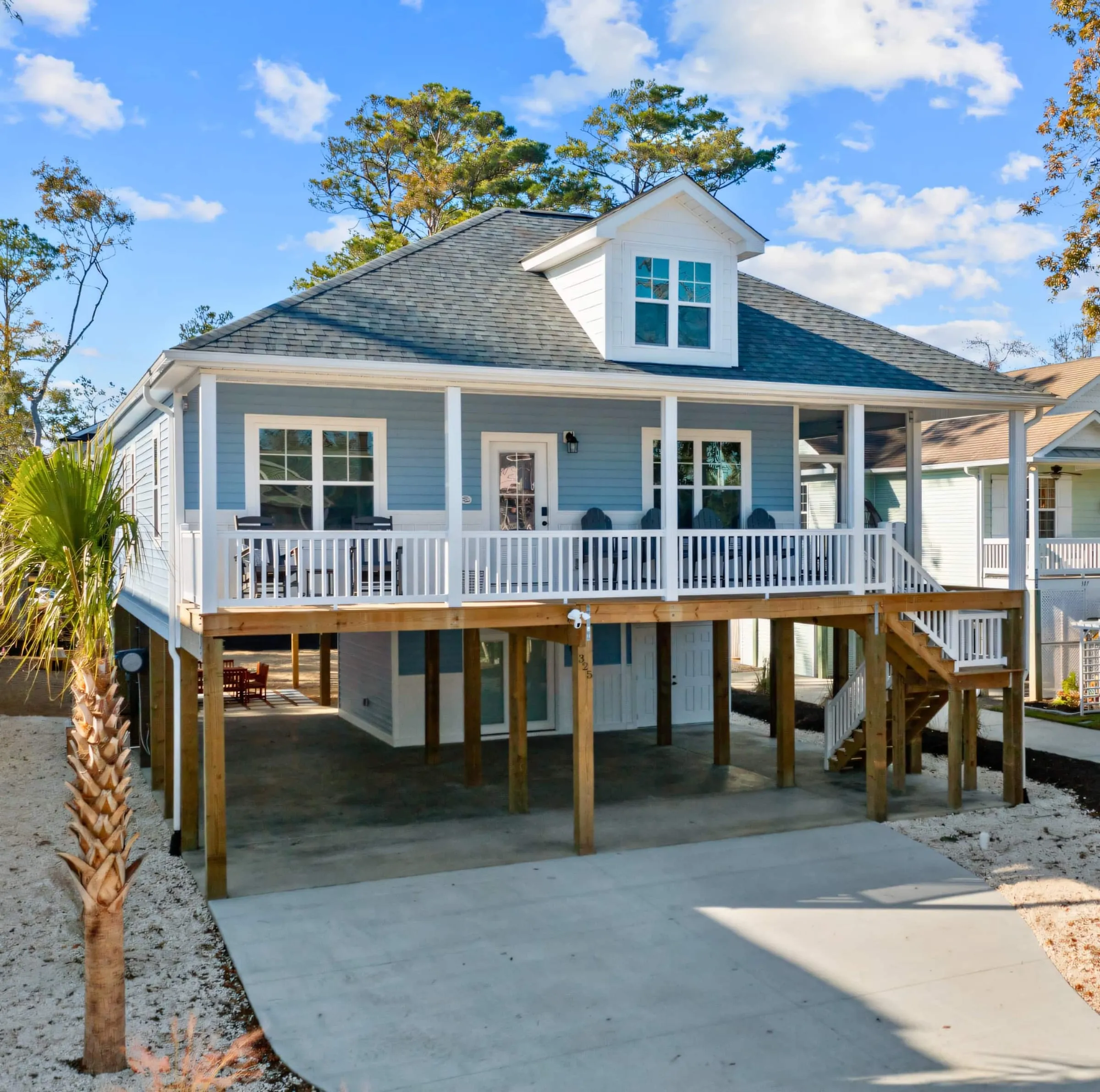 Blue Crab Beach House from the drive: raised blue cottage with a wraparound porch, a palm by the stairs, and parking underneath