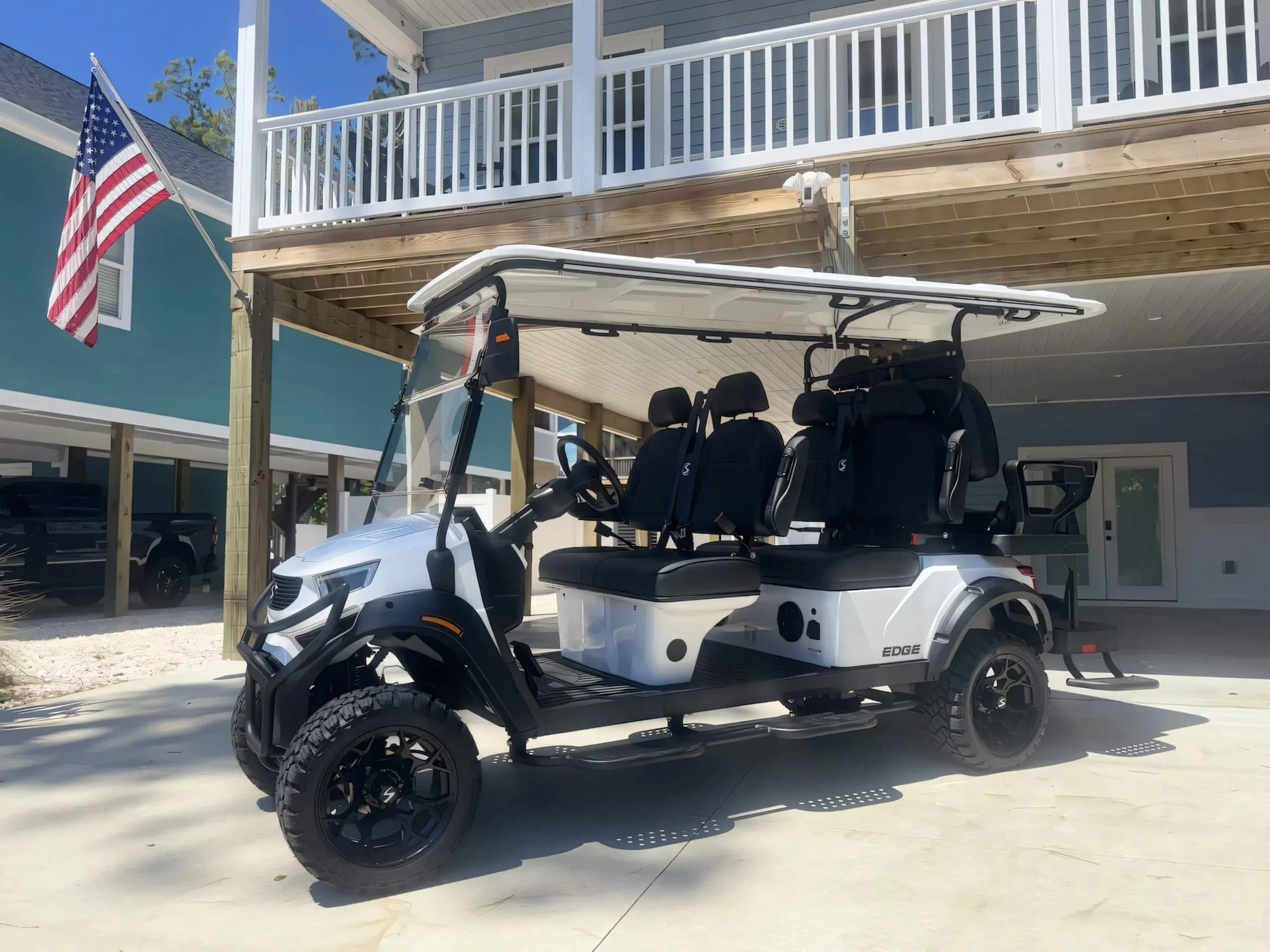 Six-seat golf cart parked under the house beside the stairs, American flag on the porch above